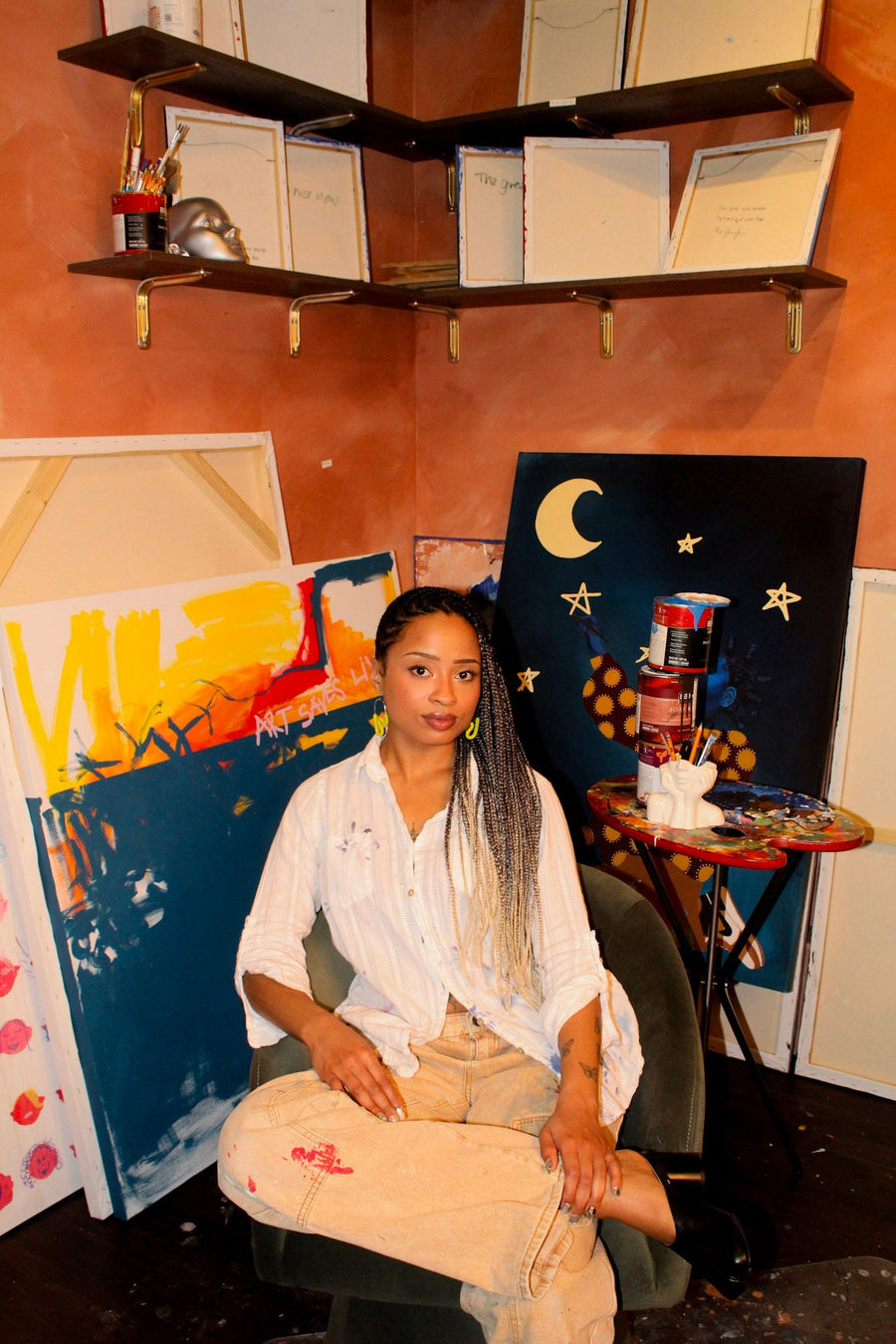 Jonarra sitting in front of pieces of her work, and paint cans to her right. 